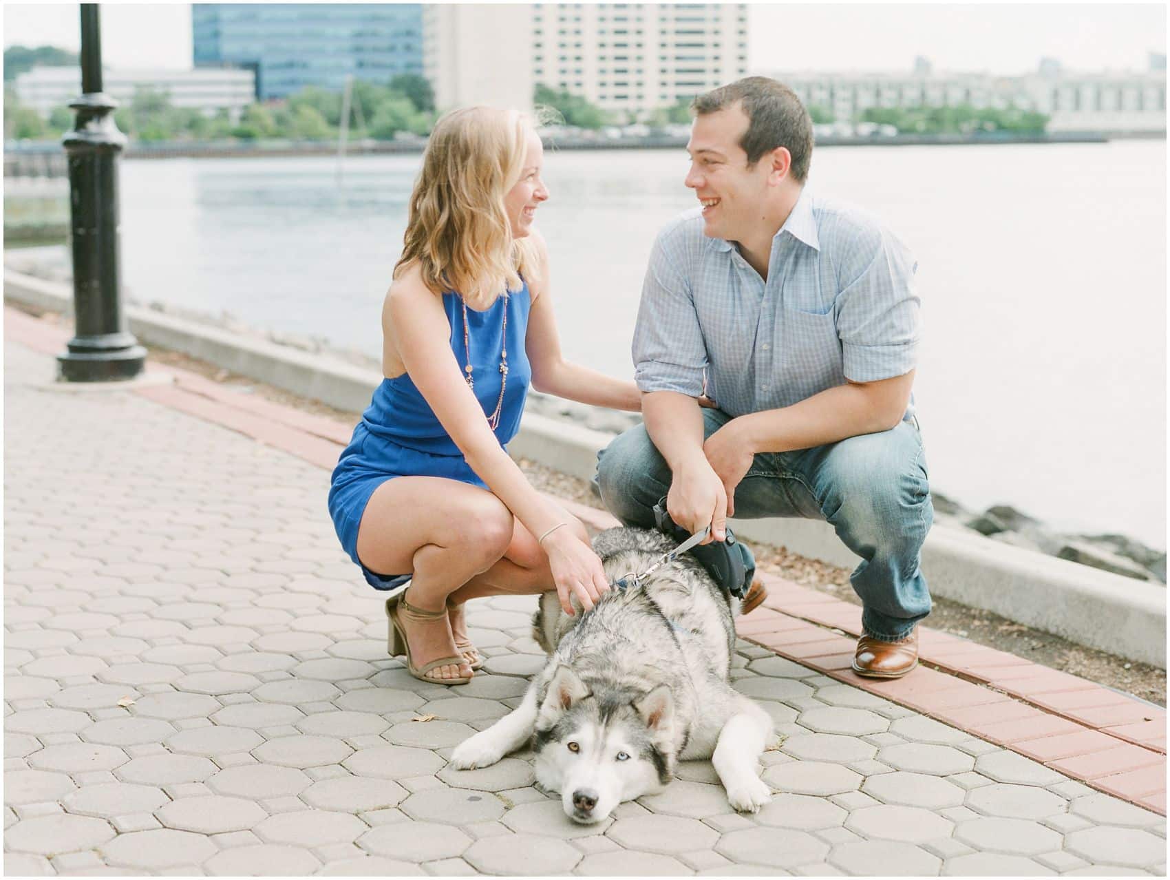 a beautiful couple with a family dog looking at each other by Hoboken Waterfront overlooking NYC