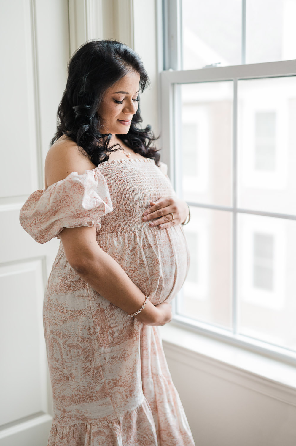 Pregnant woman in profile by window, hand on belly, floral toile maternity dress