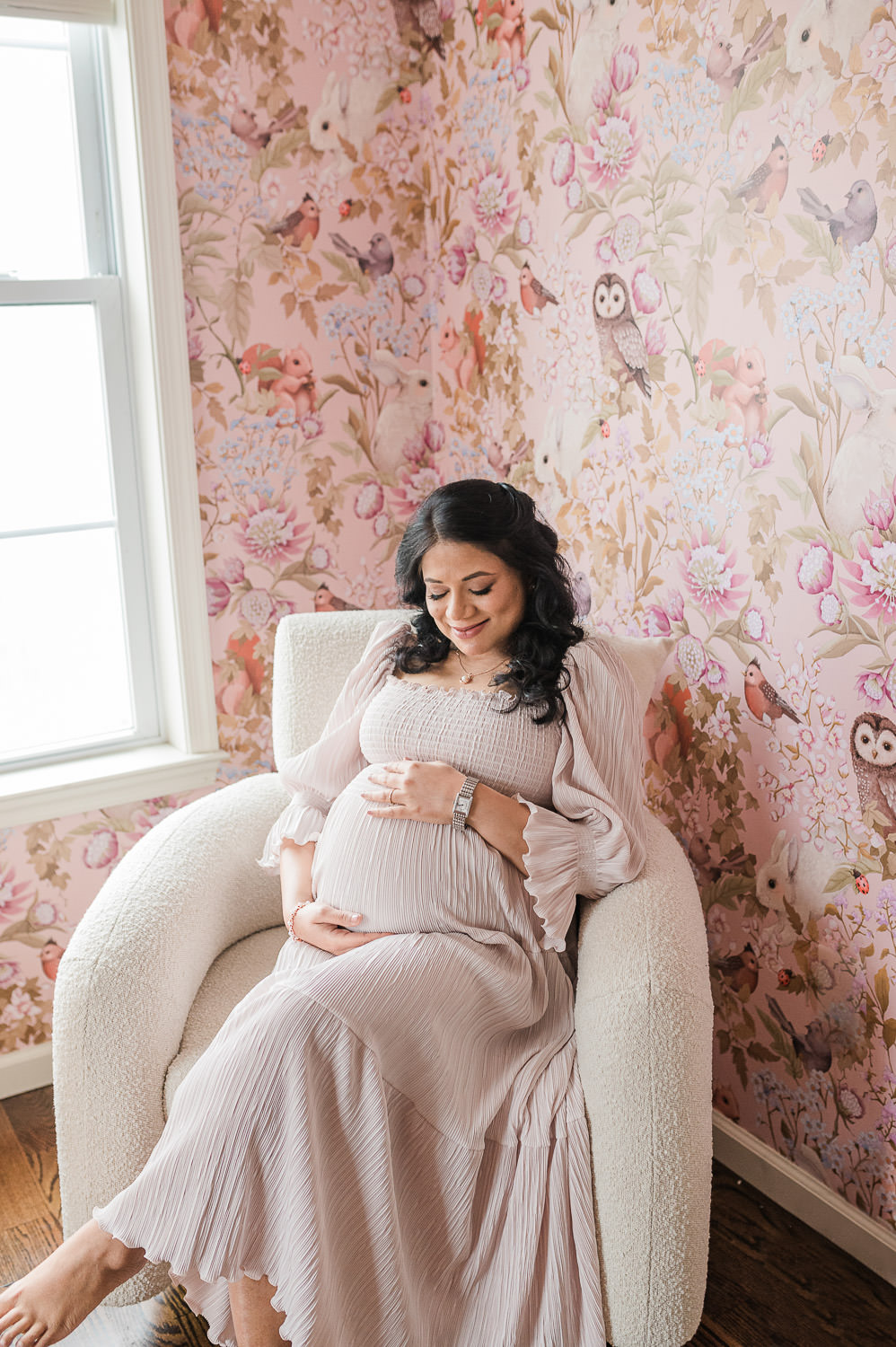 Pregnant woman seated in cream chair against pink floral wallpaper, holding belly in Hoboken Nj home