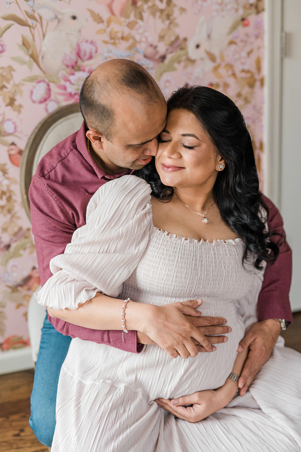 Husband embracing pregnant wife from behind, floral wallpaper nursery, hands on belly