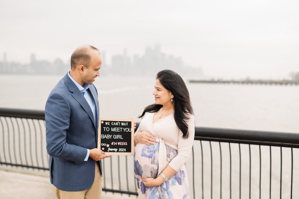 Couple at NYC waterfront holding baby announcement sign with Manhattan skyline