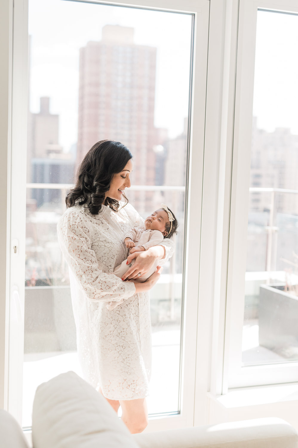 Mother holding newborn baby girl by floor to ceiling window with NYC skyline — newborn photographer Upper East Side NYC
