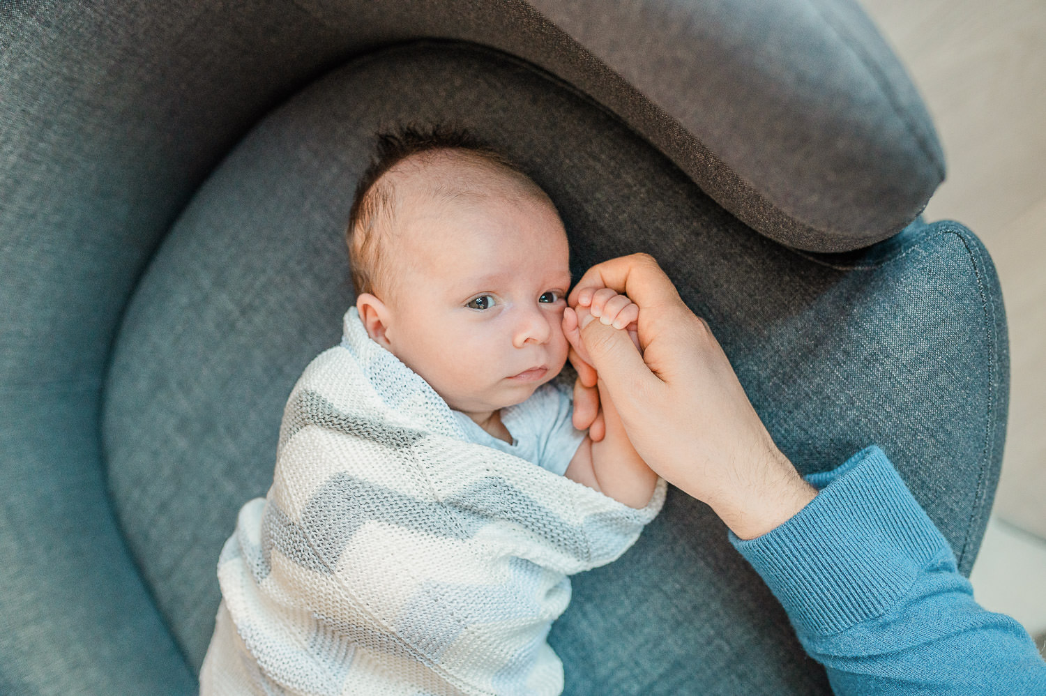 Awake newborn baby holding parent's finger in grey armchair