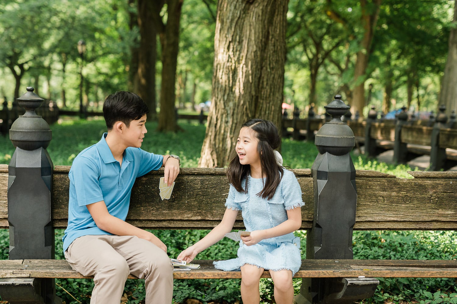 Relaxed family portrait of siblings playing cards on Central Park bench during professional photography session NYC