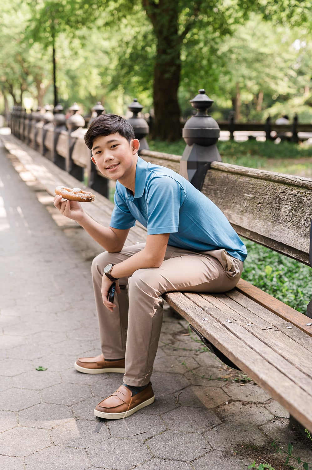 Relaxed teenage portrait of boy sitting eating pretzel on Central Park bench during professional photography session NYC