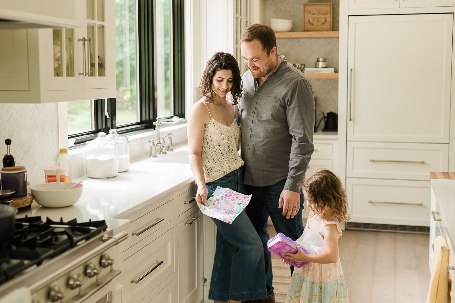 Toddler daughter proudly showing her project to happy parents by kitchen window in morning light, Summit, NJ family photography 