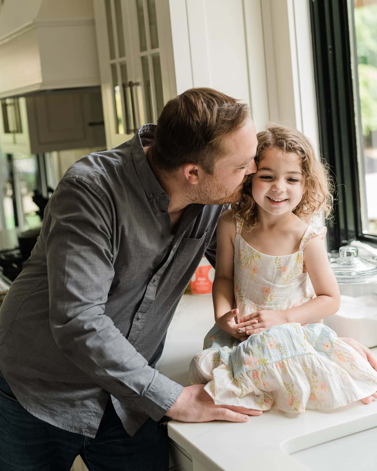 Dad kissing toddler daughter by kitchen window in morning light, Summit, NJ family photography