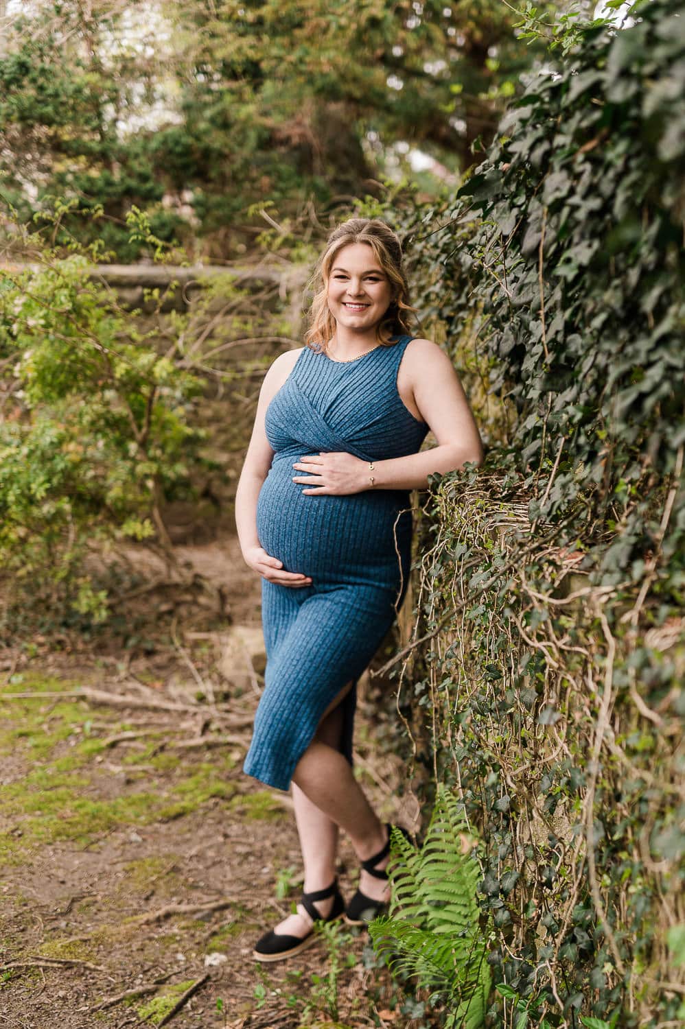Outdoor maternity photo in Morristown NJ of pregnant woman in fitted blue dress holding baby bump near ivy-covered wall