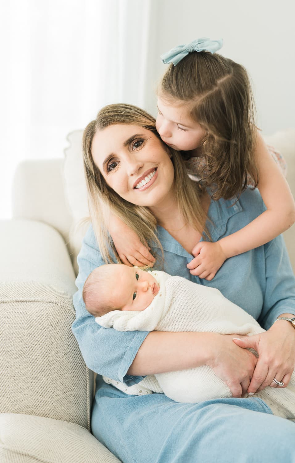 Mom in soft blue outfit holding newborn baby while older daughter kisses her cheek — lifestyle newborn photographer Short Hills NJ