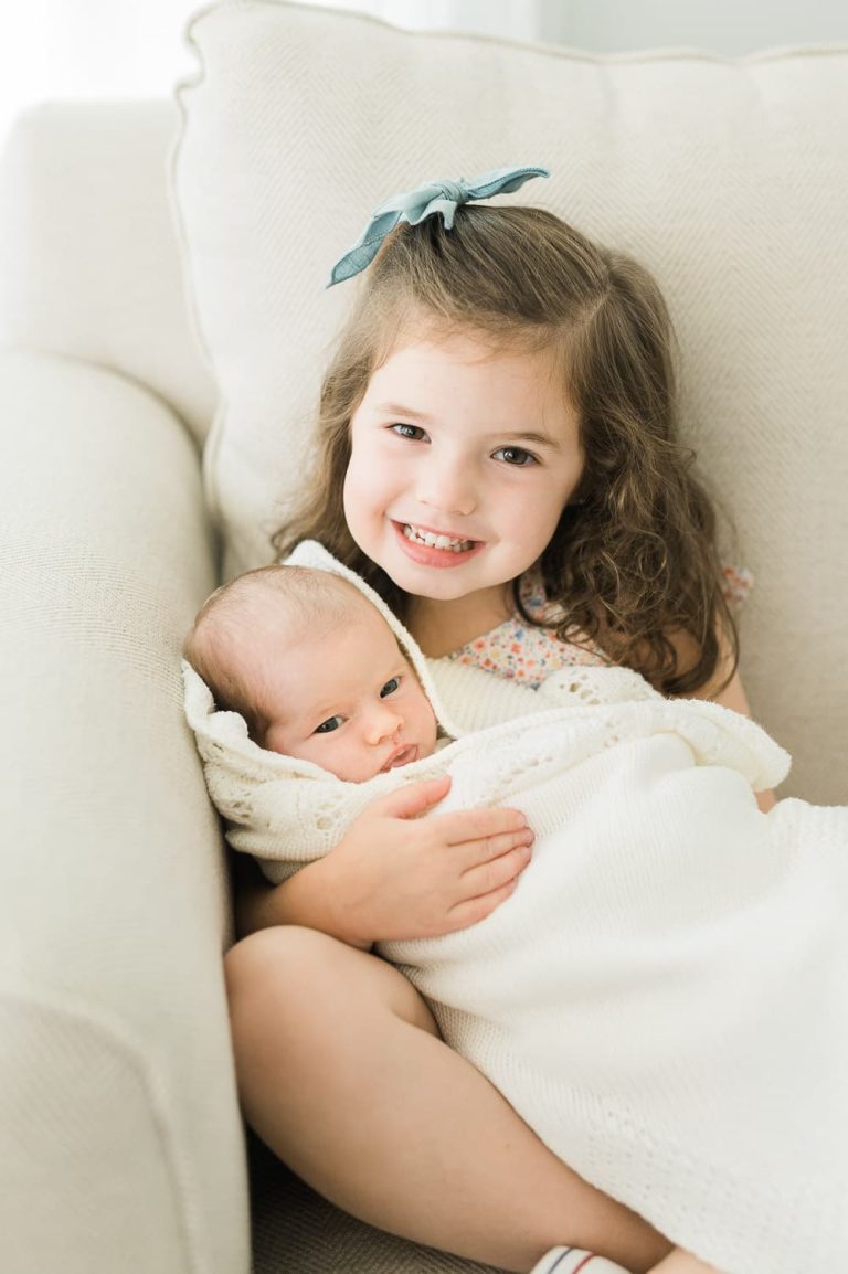 Older sister holding newborn baby sister on cream sofa — newborn photographer Short Hills NJ