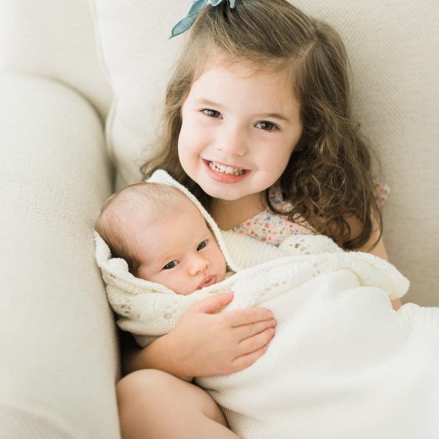 Older sister holding newborn baby sister on cream sofa — newborn photographer Short Hills NJ