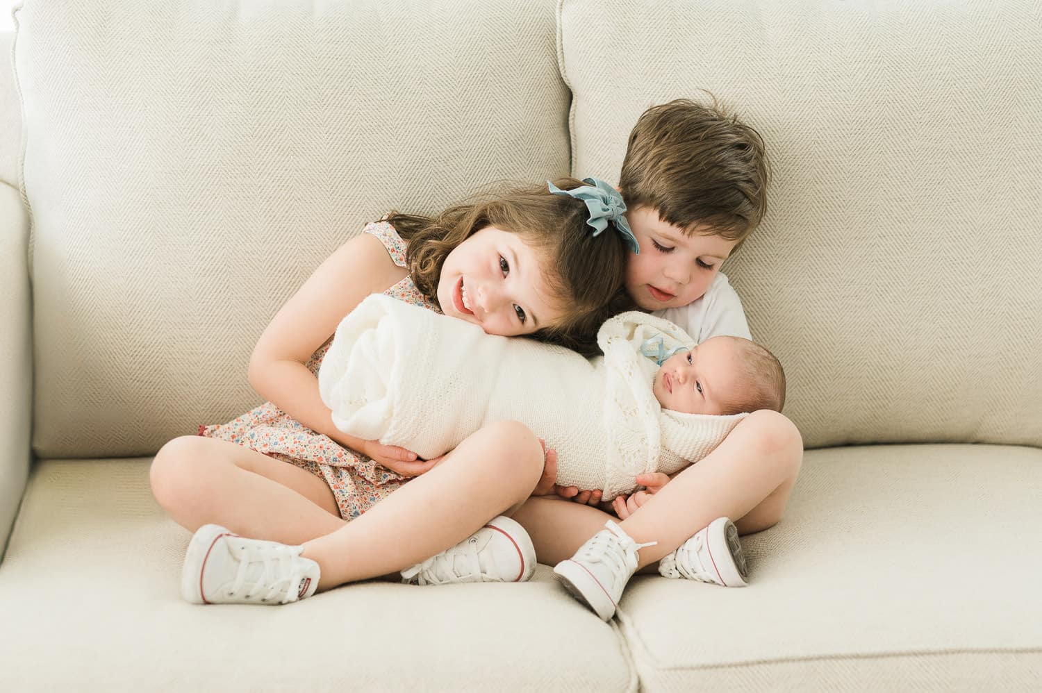 Older sister and brother cuddling newborn baby sister on a cream sofa — in-home newborn photographer Short Hills NJ