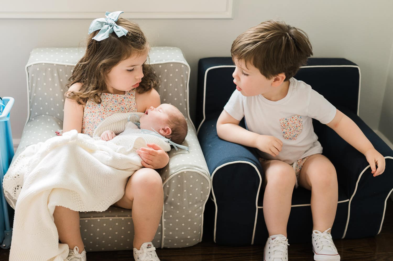 Older sister and brother sitting in their playroom chairs holding newborn baby sister — in-home newborn photography photos with Miriam Dubinsky