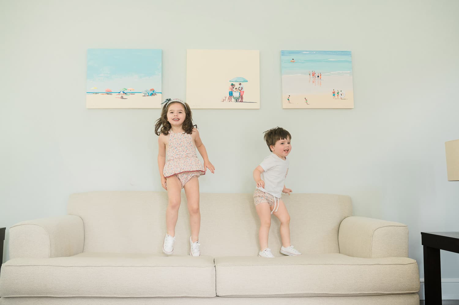 Older sister and brother jumping joyfully on a sofa in white outfits — in-home newborn photographer Short Hills NJ