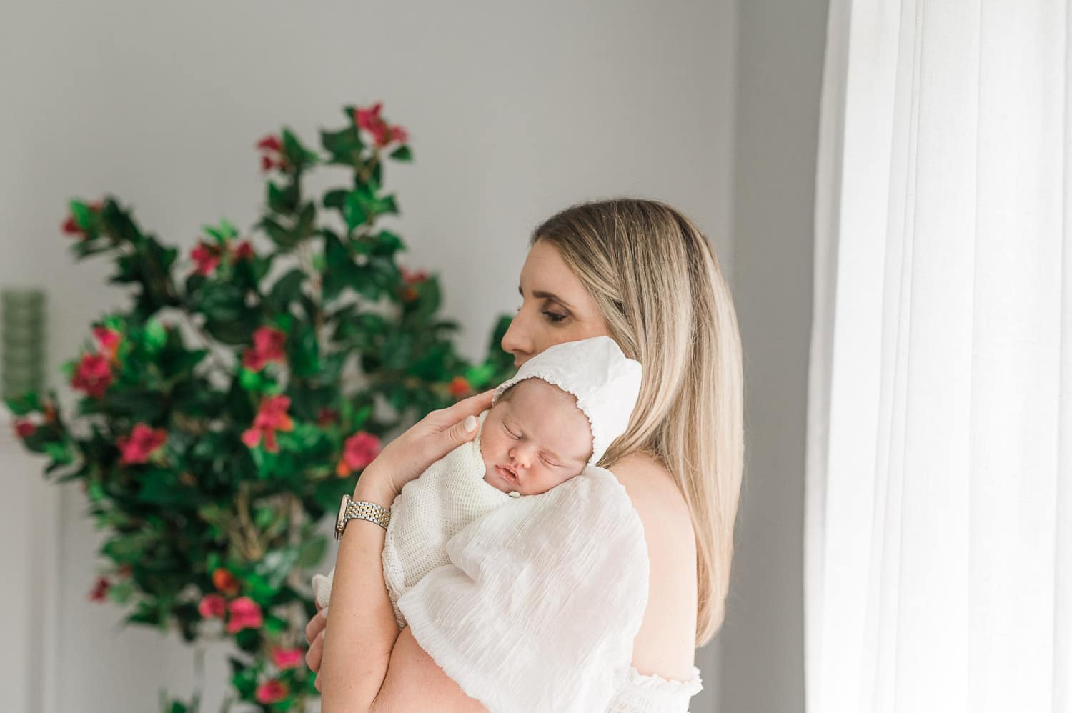 Mother holding sleeping newborn baby in white bonnet with flowers in background — at her home in Short Hills NJ