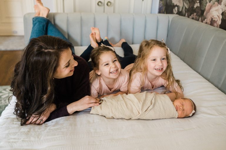 Mom giggling in bed with her three girls, including their new baby sister — newborn photos with older siblings NJ